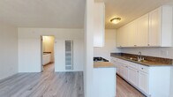 This single-floor residence offers a clean, modern aesthetic highlighted by light wood-style flooring that flows seamlessly throughout the entire space. The kitchen serves as a functional centerpiece, featuring crisp white shaker-style cabinetry, granite-look countertops, a gas cooktop, a stainless steel sink, and ample upper and lower storage. The layout includes a peninsula-style counter that helps define the kitchen area while maintaining an open connection to the adjoining living space. The living and bedroom areas are generously proportioned, bathed in natural light from large windows dressed with vertical blinds, and finished with the same consistent light flooring that gives the space a cohesive, airy feel. Vaulted ceiling angles in portions of the space add architectural interest and a sense of openness. The bathroom is well-appointed with a granite-topped vanity, white cabinetry, a large mirror, and a full tub-shower combo with a bright window above for natural light. A built-in wall vent provides climate control throughout. The overall palette of whites and neutrals creates a bright, clean environment that is both practical and inviting, making this a comfortable and move-in ready single-level home.