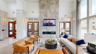This elegantly designed single-floor amenity lounge offers a generously scaled open layout that makes a strong architectural impression from the moment you enter. The defining feature of the main lounge is a dramatic floor-to-ceiling stone tile fireplace wall, clad in large-format gray marble-look tiles, which anchors the room with both warmth and grandeur. A wall-mounted flat-screen television is integrated directly above the linear fireplace, creating a refined focal point for the seating area below.

Soaring ceilings amplify the sense of openness, while an expansive wall of tall windows floods the interior with natural light and frames sweeping outdoor views of a pool and landscaped grounds. Warm-toned leather accent chairs, a neutral sofa with navy and rust cushions, and a round dark wood coffee table compose a welcoming and stylish seating arrangement.

The entry corridor features elegant herringbone-patterned tile flooring, crisp white columns, and warm wood-framed glass doors, complemented by gold-toned wall sconces throughout. A secondary kitchen area includes tall gray shaker cabinetry, stainless steel appliances, white quartz countertops, subway tile backsplash, and pendant lighting, offering a fully functional and polished space for gatherings.