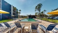 Located in Port Charlotte, Florida, this outdoor pool area offers a well-appointed recreational amenity under the sunny Southwest Florida sky. The centerpiece is a generously sized rectangular swimming pool with clear, blue-green water, blue tile accents along the edges, and lane markers indicating graduated depths — from 3 feet 6 inches on the shallow end to 4 feet toward the deeper section. Dual handrail entry points provide easy access into the water.

Surrounding the pool is a broad, paved deck finished in warm-toned interlocking tiles, providing ample space for sunbathing and relaxation. The deck is furnished with multiple sling-style lounge chairs and small side tables, thoughtfully arranged for comfort and poolside convenience. A shaded seating area with a yellow umbrella offers a retreat from the sun on one side of the deck.

At the far end of the pool, a covered pavilion with a dark roof provides additional shaded seating, ideal for gathering or outdoor dining. The entire pool area is enclosed by a black metal fence, ensuring safety and privacy. Tall palm trees and mature shade trees frame the perimeter, contributing to the tropical ambiance. The adjacent multi-story building with bold blue exterior adds a vibrant architectural backdrop to this inviting outdoor retreat.