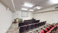 Located at 1301 W Whitestone Blvd in Cedar Park, TX, this single-floor presentation and screening room is purpose-built for gatherings, lectures, and multimedia events. The room is configured with multiple rows of theater-style seats featuring red upholstery, dark frames, and built-in cup holders, providing comfortable, audience-oriented seating for a sizeable group. A ceiling-mounted projector aligns with a large wall-mounted projection screen positioned at the front of the room, creating a clear sightline from every seat. Recessed fluorescent lighting and a textured ceiling provide even, functional illumination throughout the space. Wall-mounted speakers support audio delivery for presentations or screenings. At the front, a wooden podium stands ready for speakers, complemented by decorative potted plants that add a touch of warmth to the room. Along the rear wall, a built-in wood cabinet with countertop provides storage and surface space, accompanied by additional seating. Two entry doors offer convenient access points, and an illuminated exit sign ensures safety compliance. The patterned carpet tiles throughout add visual texture to the room. Overall, the space is well-equipped and efficiently designed for professional presentations, community screenings, or educational programming.
