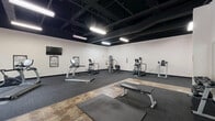Located at 44 Market Plaza in downtown Montgomery, Alabama, this single-floor fitness facility offers a well-appointed exercise space designed for a comprehensive workout experience. The open industrial-style interior features exposed black ceiling beams and ductwork, paired with bright overhead lighting and crisp white walls, creating a clean, modern atmosphere throughout.

The main exercise area is generously sized and equipped with a range of cardio machines, including treadmills and ellipticals, as well as a multi-station cable machine and a dumbbell rack with free weights. Workout posters and framed athletic photography decorate the walls, adding a motivating ambiance. Rubber flooring and gym mats provide functional workout surfaces throughout the space.

A dedicated recreation zone accommodates a full-size ping pong table, offering a casual counterpoint to the structured fitness equipment nearby. A separate lounge or transition area near the entrance features floor-to-ceiling glass walls and large windows that overlook the streetscape, flooding the space with natural light and providing open views of the surrounding urban environment. High-top seating in this area creates an inviting spot to unwind.

Additional amenities include private rooms accessible from the main floor, workout reference charts, and a welcoming, well-maintained environment suited for a variety of fitness activities.