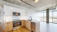 This modern 1-bedroom, 1-bathroom unit showcases an industrial-chic aesthetic defined by exposed concrete ceilings with visible ductwork, light hardwood flooring, and expansive floor-to-ceiling windows that bathe the space in natural light and frame sweeping panoramic city views. The open-concept living area seamlessly integrates the kitchen, dining, and living spaces into one cohesive layout. The kitchen is well-appointed with a quartz peninsula countertop, a marble-veined tile backsplash, light wood lower cabinetry, crisp white upper cabinets, and a full suite of stainless steel appliances including a refrigerator, electric range, microwave, and dishwasher. Matte black fixtures throughout add a refined, contemporary accent. The bedroom is generously proportioned, featuring exposed concrete columns and floor-to-ceiling windows offering elevated city vistas, creating an airy, loft-like atmosphere. Two closets provide ample storage. The bathroom is finished with large-format stone-look floor tiles, a glass-enclosed walk-in shower with white subway tile, a floating wood vanity with a white quartz countertop, matte black hardware, and a window framing additional city views. The unit's design strikes a compelling balance between industrial character and modern refinement.