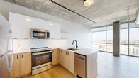 This modern 1-bedroom, 1-bathroom unit showcases an industrial-chic aesthetic defined by exposed concrete ceilings with visible ductwork, light hardwood flooring, and expansive floor-to-ceiling windows that bathe the space in natural light and frame sweeping panoramic city views. The open-concept living area seamlessly integrates the kitchen, dining, and living spaces into one cohesive layout. The kitchen is well-appointed with a quartz peninsula countertop, a marble-veined tile backsplash, light wood lower cabinetry, crisp white upper cabinets, and a full suite of stainless steel appliances including a refrigerator, electric range, microwave, and dishwasher. Matte black fixtures throughout add a refined, contemporary accent. The bedroom is generously proportioned, featuring exposed concrete columns and floor-to-ceiling windows offering elevated city vistas, creating an airy, loft-like atmosphere. Two closets provide ample storage. The bathroom is finished with large-format stone-look floor tiles, a glass-enclosed walk-in shower with white subway tile, a floating wood vanity with a white quartz countertop, matte black hardware, and a window framing additional city views. The unit's design strikes a compelling balance between industrial character and modern refinement.