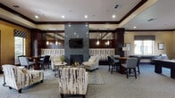 This well-appointed single-floor community space combines upscale design with practical amenities, creating an inviting environment for relaxation and social interaction. The centerpiece of the space is a generously sized lounge area anchored by a striking floor-to-ceiling stone fireplace wall with a mounted flat-screen television, surrounded by carefully arranged seating including patterned accent chairs, sofas, and intimate groupings set atop geometric-patterned area rugs. Rich dark wood trim and crown molding frame the warm, neutral-toned walls throughout, lending a refined and cohesive aesthetic. Pendant lighting and recessed ceiling lights provide layered illumination, enhancing the inviting ambiance. A dining area with dark wood tables and upholstered chairs is positioned near a glass door that opens to an outdoor area, allowing natural light to filter in. The kitchen is fully equipped with dark cabinetry, granite-style countertops, black appliances including a range, over-the-range microwave, and a side-by-side refrigerator with water and ice dispenser, along with a prep sink and ample counter space. A secondary lounge area features comfortable seating, a wall-mounted television, and access to a glass-door office or management suite. The overall atmosphere is polished and welcoming, suited for community gatherings and everyday use.