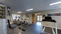 This Fitness and Business Center is a well-appointed, single-floor facility designed to serve the health and professional needs of its users. The fitness area is outfitted with a variety of cardio equipment, including an elliptical trainer, stationary spin bikes, and a treadmill, all arranged on durable rubber flooring to support a safe and comfortable workout environment. Recessed fluorescent lighting and windows with blinds provide a bright, well-lit atmosphere throughout the space. Neutral-toned walls give the interior a clean and focused feel. A transition through an open doorway leads into a connected area featuring hardwood-style flooring, which flows toward a set of glass entry doors that welcome in natural light and offer views of the surrounding outdoor greenery. This passage connects the fitness zone to the business center portion of the facility, creating a seamless, multi-purpose space. Wall-mounted accessories and organized equipment placement reflect a thoughtfully arranged environment built for both functionality and ease of use. The combination of fitness and business amenities within a single accessible location makes this center a practical and versatile resource.