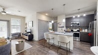 This thoughtfully designed one-bedroom unit offers an efficient and stylish layout across a single floor. The open-concept living and kitchen area serves as the heart of the space, featuring rich wood-style flooring that flows seamlessly throughout. The kitchen is equipped with dark charcoal cabinetry, a mosaic tile backsplash, stainless steel appliances—including a refrigerator, range, and microwave—and a granite-topped peninsula with bar seating illuminated by pendant lighting. A dedicated dining nook adjacent to the kitchen provides an intimate space for meals. The living room is bright and welcoming, with large windows allowing natural light to fill the room, complemented by a ceiling fan overhead. A set of double doors leads from the main living area toward the bedroom wing. The generously sized primary bedroom features plush carpeting, a ceiling fan, and a large window that brings in ample natural light. It connects to a walk-in closet outfitted with wire shelving systems on multiple walls, offering substantial storage capacity. Throughout the unit, neutral wall tones and consistent finishes create a cohesive, modern aesthetic that balances comfort with contemporary design.