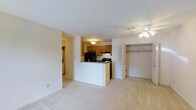 This single-floor space offers a practical and comfortable layout designed for everyday living. The main living area is open and airy, finished with light-toned wall-to-wall carpeting that extends throughout the space, creating a warm and cohesive feel. A ceiling fan with integrated lighting provides both comfort and illumination to the living area, while a generously sized closet with built-in shelving and a bifold door offers ample storage directly off the main room.

The kitchen is efficiently laid out and partially open to the living space via a raised breakfast bar, which serves as both a functional workspace and a casual dining surface. The kitchen features warm wood-toned cabinetry, dark countertops, and black appliances including a refrigerator and range with overhead ventilation. A flush-mount ceiling light brightens the cooking area. Adjacent to the kitchen, a hallway connects to additional areas of the space, hinting at a well-organized floor plan. The textured ceilings and neutral wall tones throughout contribute to a clean, understated aesthetic that provides a versatile backdrop for a variety of interior styles.
