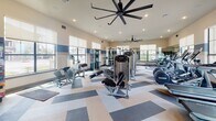 Located at 21201 Emerald Mist Pkwy in Spring, TX, this fitness center is a single-floor facility designed to support a comprehensive workout experience. The space is generously sized, featuring large windows along multiple walls that flood the interior with natural light and offer views of the surrounding community. Roller shades provide flexible light control throughout the day.

The flooring features a bold geometric pattern in shades of blue, gray, and tan, adding a dynamic visual element to the space. Overhead, large ceiling fans with a modern industrial design ensure consistent air circulation throughout the facility.

The strength training area is well-appointed with a variety of cable machines, multi-functional weight stations, and Precor-branded equipment suited for both isolated and compound exercises. A dedicated functional training zone includes a vertical rack stocked with kettlebells, medicine balls, and resistance bands, supporting a wide range of movement-based workouts. Wall-mounted mirrors expand the sense of space and allow users to monitor their form.

The cardio section offers an array of equipment including treadmills, ellipticals, stationary bikes, and recumbent cycles. A dumbbell rack with a full range of free weights rounds out the offerings. Recessed lighting and accent walls featuring bold navy blue horizontal stripes contribute to the facility's polished, motivating atmosphere.