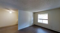 This single-floor residence offers a straightforward and functional layout designed for comfortable everyday living. The main living area is finished with rich, dark hardwood flooring that contrasts cleanly against the neutral white walls, creating a simple yet classic aesthetic. A large window allows natural light to fill the room, brightening the space and offering a view of the outdoors. The textured popcorn ceiling adds a layer of traditional character overhead. A baseboard heating unit runs along the exterior wall beneath the window, providing an efficient and unobtrusive heat source. An open entryway connects the main living area to an adjacent room or hallway, suggesting a practical, open-feeling floor plan. The walls are painted in a soft, neutral tone throughout, lending the space a clean and versatile canvas suitable for a variety of interior styles. Overall, the space presents a functional and understated residential environment with key features that prioritize practicality and ease of living.