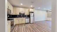 This single-floor unit presents a cohesive modern design characterized by warm wood-look luxury vinyl plank flooring throughout the main living areas and durable carpet in the bedrooms. The kitchen is a standout feature, outfitted with light maple cabinetry, matte black hardware, a bold black subway tile backsplash, and a full suite of stainless steel appliances including a refrigerator, dishwasher, range, and over-the-range microwave. Recessed lighting illuminates the space efficiently throughout.

The open living area connects seamlessly to the kitchen and features sliding glass doors that open to a patio, allowing natural light to filter in and providing outdoor access. Exposed silver ductwork lends an industrial accent to certain areas of the unit.

The bedrooms are carpeted for comfort, with one room featuring a window with an outdoor view and another accessed through a distinctive sliding barn door — a stylish architectural detail that adds character. The bathroom features matching light maple cabinetry, a vanity with a large mirror, and consistent luxury vinyl plank flooring, maintaining design cohesion throughout the unit.