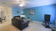 This two-story space makes a strong visual impression with its living area characterized by vibrant teal-painted walls that create a warm, lively atmosphere. The living room is generously sized, offering ample space for comfortable furnishings, including a leather sofa and a glass-top coffee table that anchor the room. Soft, neutral carpeting runs throughout, providing a comfortable contrast to the bold wall color. A ceiling fan with an integrated light fixture is centrally positioned, ensuring both comfort and adequate lighting. Decorative wall art adds personality to the space, complementing the overall aesthetic. The room benefits from good proportions, making it well-suited for both relaxing and entertaining. As a two-story property, residents can expect additional living, sleeping, or functional spaces across both levels, offering a practical separation between common and private areas. The overall ambiance of the living area is casual and inviting, with the distinctive teal walls serving as a defining design feature throughout.