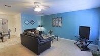 This two-story space makes a strong visual impression with its living area characterized by vibrant teal-painted walls that create a warm, lively atmosphere. The living room is generously sized, offering ample space for comfortable furnishings, including a leather sofa and a glass-top coffee table that anchor the room. Soft, neutral carpeting runs throughout, providing a comfortable contrast to the bold wall color. A ceiling fan with an integrated light fixture is centrally positioned, ensuring both comfort and adequate lighting. Decorative wall art adds personality to the space, complementing the overall aesthetic. The room benefits from good proportions, making it well-suited for both relaxing and entertaining. As a two-story property, residents can expect additional living, sleeping, or functional spaces across both levels, offering a practical separation between common and private areas. The overall ambiance of the living area is casual and inviting, with the distinctive teal walls serving as a defining design feature throughout.