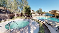 Located at 12963 Moreno Beach Dr in Moreno Valley, CA, this outdoor amenity space offers a resort-style retreat designed for relaxation and recreation. The centerpiece is a generously sized swimming pool with crystal-clear blue water, complemented by a separate spa/hot tub, providing options for both active swimming and leisurely soaking. The expansive concrete pool deck is lined with multiple lounge chairs and shaded by teal market umbrellas, creating inviting spots to enjoy the Southern California sunshine.

A covered pergola structure with rich wood beams and stone-accented columns anchors the outdoor kitchen area, which features dual stainless steel built-in gas grills set into a stone masonry countertop — ideal for community gatherings and casual outdoor dining. Mature trees and lush landscaping surround the perimeter, offering natural shade and a sense of privacy. Decorative stone walls and wrought iron fencing define the space while maintaining open sightlines to the surrounding hillside and multi-story residential buildings. The overall design blends Craftsman-inspired architectural details with functional outdoor living, creating a well-appointed communal environment suited to the warm Inland Empire climate.