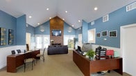 This single-floor space serves as a well-appointed leasing or community office, thoughtfully designed to be both functional and welcoming. The most commanding architectural feature is the soaring vaulted ceiling, which rises dramatically toward a central peak and is lined with recessed lighting that evenly illuminates the entire interior. A full-height brick chimney anchors the room, serving as a bold focal point with a wall-mounted flat-screen television positioned above the fireplace structure.

The walls are finished in a warm, muted blue tone accented by white wainscoting trim, creating a clean and professional atmosphere. The space is divided into two primary functional areas: a comfortable lounge seating area featuring a dark leather sofa arranged facing the fireplace, and a dedicated reception and work station equipped with a wooden desk, computer, printer, and organized paperwork trays.

Natural light filters in generously through multiple sets of French-style double doors and windows on both sides of the room, enhancing the open and airy feel. Decorative touches such as potted plants and framed artwork add warmth to the professional setting. Neutral carpeting throughout ties the space together, while the overall layout ensures a smooth and accessible flow between the reception and seating zones.