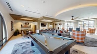 Located at 1625 Santa Venetia St in Chula Vista, CA, this single-floor community clubhouse is thoughtfully designed to offer a variety of social and recreational spaces. The open-plan layout flows seamlessly between zones, unified by a contemporary design palette of warm wood tones, stone countertops, and decorative tiled flooring.

The game and lounge area serves as a central gathering point, featuring a full-size pool table set atop a graphic area rug, along with a wet bar complete with bar seating and a wall-mounted TV. Distinctive circular tray ceiling details with ambient cove lighting add architectural character throughout the space.

The kitchen and dining area is equipped with stainless steel appliances, an expansive marble-topped island, warm wood cabinetry, and a textured rope-screen partition that adds a natural, artisanal element. Multiple dining tables and mixed seating arrangements make this area ideal for group gatherings.

A comfortable lounge area features a herringbone wood accent wall with a large flat-screen TV, a plush sectional sofa, and an assortment of accent chairs, all oriented around floor-to-ceiling steel-framed windows that invite abundant natural light and outdoor views.

A separate media and relaxation room offers an oversized navy sectional sofa, bean bag seating, and a tray ceiling with cove lighting, providing a more intimate and casual setting for entertainment.