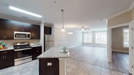 This well-appointed single-floor residence offers a thoughtfully designed open-concept layout that seamlessly connects the kitchen, dining, and living areas. The kitchen is a standout feature, showcasing rich dark espresso cabinetry, granite countertops, and a full suite of stainless steel appliances including a refrigerator, range, microwave, and dishwasher. A central kitchen island with pendant lighting provides additional prep space and casual seating. Crown molding elegantly frames the main living and dining areas, which are bathed in natural light from large windows with exterior views. Light hardwood-style flooring flows throughout the living spaces, complemented by neutral, warm-toned walls that create a bright and welcoming ambiance. The living and dining rooms are further enhanced by ceiling fans and decorative chandeliers. The bedroom features a pendant light fixture, a large window, and the same hardwood-style flooring. The bathroom includes a dark-cabineted vanity with granite countertops, tile flooring, and access to a walk-in closet area. Overall, the space presents a clean, modern aesthetic with quality finishes throughout.