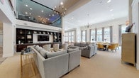 This expansive single-floor clubhouse is designed to accommodate a variety of social and functional needs, combining elegant design with practical amenities across several distinct zones. The main lounge area commands attention with its dramatically high ceilings, a mezzanine-level overlook with glass railings, and a pair of ornate chandeliers that anchor the space. Multiple sectional sofas in neutral grey tones are arranged around a built-in entertainment wall featuring dark wood shelving and a large flat-screen television, creating an inviting gathering area.

Adjacent to the lounge, a bright casual seating area features floor-to-ceiling windows that flood the space with natural light and offer views of the surrounding community. A gold chandelier and hardwood-style flooring add warmth, while round dining tables with teal-accented chairs provide flexible seating. A formal private dining room includes a large herringbone-topped wood table, tufted upholstered chairs, and an antler chandelier, accessible via glass-paneled doors.

The full catering kitchen is outfitted with dark espresso cabinetry, granite countertops, a tile backsplash, stainless steel appliances, and a center island with bar seating. Throughout the clubhouse, a cohesive palette of soft neutrals, warm wood tones, and metallic accents creates a polished and welcoming ambiance.