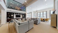 This expansive single-floor clubhouse is designed to accommodate a variety of social and functional needs, combining elegant design with practical amenities across several distinct zones. The main lounge area commands attention with its dramatically high ceilings, a mezzanine-level overlook with glass railings, and a pair of ornate chandeliers that anchor the space. Multiple sectional sofas in neutral grey tones are arranged around a built-in entertainment wall featuring dark wood shelving and a large flat-screen television, creating an inviting gathering area.

Adjacent to the lounge, a bright casual seating area features floor-to-ceiling windows that flood the space with natural light and offer views of the surrounding community. A gold chandelier and hardwood-style flooring add warmth, while round dining tables with teal-accented chairs provide flexible seating. A formal private dining room includes a large herringbone-topped wood table, tufted upholstered chairs, and an antler chandelier, accessible via glass-paneled doors.

The full catering kitchen is outfitted with dark espresso cabinetry, granite countertops, a tile backsplash, stainless steel appliances, and a center island with bar seating. Throughout the clubhouse, a cohesive palette of soft neutrals, warm wood tones, and metallic accents creates a polished and welcoming ambiance.