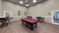 This single-floor game room is thoughtfully designed as a dedicated recreational space, combining functional amenities with a comfortable, inviting atmosphere. The room's centerpiece is a full-size pool table with a rich red felt surface and ornate dark wood legs, providing ample space around it for gameplay. The room's generous ceiling height, complemented by a drop ceiling with recessed panel lighting, creates a bright and open feel throughout.

The flooring consists of warm-toned hardwood-style planks that run the length of the space, adding character and durability. A distinctive white chair rail runs along the perimeter of the walls, dividing the neutral beige upper walls from the crisp lower wainscoting, lending the room a polished, finished look.

Framed artwork adorns the walls, adding visual interest without overwhelming the space. Bar-height bistro tables paired with swivel stools are positioned along the walls, offering casual seating for spectators or social gatherings. A wall-mounted flat-screen television provides an additional entertainment option.

Two doorways provide access to and from the space, one featuring a glass-panel insert. The overall ambiance is relaxed yet refined, making it a versatile setting for leisure and community engagement.