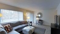 This single-floor space presents a welcoming living area characterized by a clean, neutral aesthetic and comfortable layout. The room is anchored by plush wall-to-wall carpeting in a warm beige tone, contributing to a cozy atmosphere throughout. A large window dressed with vertical blinds and sheer curtains allows generous natural light to fill the space, creating an airy, open feel.

The living area is thoughtfully arranged, featuring a spacious sectional sofa in soft grey, complemented by bold chevron-patterned accent pillows that add a touch of visual interest. A tufted ottoman serves as a central coffee table, paired with a grey area rug that defines the seating zone. Decorative candle holders on a tray atop the ottoman add an elegant finishing detail.

Along one wall, a sleek console table supports a table lamp and small décor items, while a striking sunburst mirror mounted above serves as a notable focal point, enhancing the room's sense of depth and style. A small desk and chair tucked into the corner offer a functional workspace without disrupting the room's overall flow. The smooth, white ceiling and light-toned walls maintain a bright, cohesive ambiance throughout.