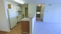 This single-floor residential unit offers a practical and functional layout designed to maximize everyday comfort. The kitchen is a standout feature, equipped with white appliances including a range and refrigerator, ample cabinetry with warm wood-toned accents, and generous counter space. A peninsula-style breakfast bar with matching wood-front paneling extends into the adjacent area, creating a natural divide between the kitchen and the living space while encouraging an open, connected feel. The kitchen flooring features wood-look hardwood-style planks that add warmth and are easy to maintain. Transitioning from the kitchen, the living area is finished with plush carpeting that runs throughout, providing comfort underfoot. A hallway leads to the remaining rooms, where built-in shelving and closet storage solutions offer organized spaces for everyday essentials. The layout benefits from overhead lighting throughout, ensuring each area is well-lit. A walk-in or reach-in closet is visible along the hallway corridor, further enhancing the storage capacity of the unit. Overall, the space presents a clean, neutral palette with warm wood accents, offering a comfortable and move-in-ready environment well-suited for practical, day-to-day living.