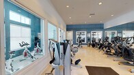 Located at 25550 River Bend Dr in Yorba Linda, California, this well-appointed fitness center offers a comprehensive, single-floor facility designed to support a wide range of workout needs. The space features light hardwood-style flooring, crisp white ceilings with recessed lighting, and striking blue accent walls that create an energetic yet clean atmosphere throughout.

The main workout floor is generously sized and lined with floor-to-ceiling mirrors along one wall, enhancing the sense of openness and allowing members to monitor their form. A robust selection of Cybex cardio equipment includes multiple treadmills, elliptical trainers, and stationary bikes arranged in organized rows. Strength training is well-represented with a multi-station cable machine, including an HD1000 functional trainer, along with various resistance and weight machines positioned throughout the space.

Large windows along the perimeter flood the facility with natural light and provide views to the surrounding landscape. A separate enclosed room, accessible via glass-paneled doors, is dedicated to indoor cycling and spin bikes. Adjacent to it is a bright, colorful children's play area, making the facility family-friendly and accessible for parents. The overall design balances functionality and comfort, offering a clean, professional environment suitable for all fitness levels.