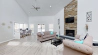 This single-floor clubhouse offers an inviting and well-appointed gathering space designed with both comfort and functionality in mind. The main living area immediately draws the eye upward to its dramatic vaulted ceiling, which creates an expansive, airy atmosphere throughout the room. A ceiling fan complements the open height, ensuring year-round comfort. Anchoring the space is a floor-to-ceiling stacked stone fireplace, a standout architectural feature that adds warmth and texture to the otherwise clean, contemporary aesthetic. A wall-mounted television above the fireplace makes the area equally suited for casual viewing or social events. The living area is furnished with plush, tufted seating arranged around a dark-toned coffee table, offering a comfortable and stylish setting for residents to relax or gather. Light hardwood-style flooring runs throughout, reflecting the abundant natural light that streams in through multiple windows. Adjacent to the main seating area, a dedicated workspace with desks and chairs provides a practical area for residents who need a quiet place to work or study. The overall palette of soft whites, neutral furnishings, and natural stone accents gives the clubhouse a modern yet welcoming character, making it an ideal communal hub for any community.