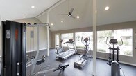 This single-floor fitness center is designed to provide a functional and comfortable workout environment. The space features vaulted ceilings with exposed beams that lend an open, airy feel, complemented by recessed lighting and a modern ceiling fan to maintain airflow during workouts. Large windows line the walls, flooding the interior with natural light and offering pleasant views of the surrounding greenery, creating an inviting atmosphere that enhances the exercise experience. The flooring is a durable, dark-toned surface well-suited for high-traffic fitness use. The space is equipped with a variety of cardio machines, including a treadmill, an elliptical trainer, and a stationary bike, catering to a range of cardiovascular workout preferences. A multi-station strength training cable machine is positioned along one wall, alongside a weight bench, providing options for resistance and strength training. Structural support columns are integrated throughout the space, and neutral-toned walls contribute to a clean, focused aesthetic. Overall, the fitness center balances practicality with a bright, welcoming design that encourages regular use.