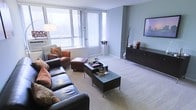 This single-floor space centers around a well-appointed living area designed for both comfort and functionality. Large floor-to-ceiling windows along one wall flood the room with abundant natural light, offering views of the surrounding outdoors while vertical blinds allow for adjustable privacy. The soft, neutral-toned carpet underfoot adds warmth and a sense of ease throughout the space. The living area is anchored by a substantial dark leather sofa and a matching leather armchair with ottoman, providing generous seating options. A set of sleek, dark-finished coffee tables with chrome legs serves as the focal point of the seating arrangement, complementing the room's modern aesthetic. A wall-mounted flat-screen television is positioned above a dark, low-profile media console, which offers additional storage. The walls are finished in a cool, muted blue-grey tone that contributes to the calm, contemporary ambiance of the space. Decorative accents, including dried branch arrangements and carefully placed accessories, add subtle visual interest without overwhelming the clean lines of the interior. The overall design balances minimalism with comfort, creating a functional and inviting living environment.