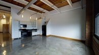 This single-floor space showcases a compelling industrial aesthetic, blending raw materials with functional design. The polished concrete floors extend throughout the open-concept layout, reflecting light and adding a sleek, modern character to the interior. Overhead, a striking wood-paneled ceiling with exposed beams and metal ductwork creates a warm contrast against the otherwise industrial elements, lending the space a loft-like ambiance with generous ceiling height.

The kitchen is positioned along one wall and is well-equipped with black appliances, including a range, oven, and refrigerator. Dark countertops run along the workspace, complemented by white cabinetry featuring frosted glass upper cabinet doors. A stainless steel sink sits beneath pendant lighting, which hangs from the ceiling in a row, providing both task lighting and visual interest.

The open living area is expansive and versatile, benefiting from the high ceilings and clean white walls that create a bright, airy atmosphere. A partial interior wall with a sloped detail adds architectural interest to the space. The warm wood baseboard trim throughout provides a subtle finishing touch, tying together the industrial and natural elements of this distinctive space.