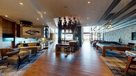 This sophisticated single-floor amenity space is designed to accommodate a variety of social and recreational activities within one expansive, open-plan layout. Wide-plank hardwood flooring flows throughout the main area, anchored by a dramatic, multi-armed sculptural chandelier suspended from a recessed ceiling detail that serves as the visual centerpiece of the room. Floor-to-ceiling windows bathe the interior in natural light while offering panoramic outdoor views, complemented by a slat-wood ceiling detail in the lounge zone.

The space is organized into distinct functional areas. A full demonstration kitchen features a substantial marble-topped island with turquoise upholstered bar seating, a gas range with a concrete hood, iridescent tile backsplash, and pendant lighting. A wet bar area mirrors this aesthetic with sleek grey cabinetry, gold hardware, a continuous light shelf, and the same distinctive tile treatment.

A games area includes a full-size billiards table, while a cozy lounge nook offers a fireplace, plush sectional seating, and a built-in bookshelf. A bar-height ledge along a stacked stone accent wall provides additional seating with a mounted television. The overall ambiance is upscale, layered, and inviting.