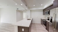 This thoughtfully designed single-floor unit presents a clean, contemporary aesthetic characterized by crisp white walls, light wood-tone flooring, and recessed lighting throughout. The open-concept kitchen serves as a central highlight, featuring an extended white quartz island with a double undermount sink and a pull-down faucet, complemented by rich dark wood cabinetry. The full appliance suite includes stainless steel refrigerator, range, and built-in microwave, all set against a bright, functional layout. The kitchen flows seamlessly into a generous living area, where high ceilings and recessed lighting create an open, airy ambiance. Bedrooms are spacious and bright, with large windows dressed in vertical blinds that allow abundant natural light to filter in. The primary bedroom connects directly to a well-organized walk-in closet outfitted with multiple chrome hanging rods and light wood flooring. The bathroom features marble-look tile flooring, a white quartz vanity countertop, dark wood cabinetry with chrome hardware, a large mirror, and clean modern fixtures. The overall design is cohesive and modern, with a neutral palette that offers a versatile canvas throughout every room.