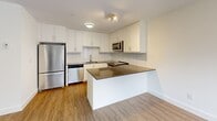 Located at 77 Parkwoods Village Dr in Toronto's Parkwoods-Donalda neighbourhood, this one-bedroom, one-bathroom unit offers a well-designed layout with modern finishes throughout. The kitchen is a standout feature, showcasing crisp white shaker-style cabinetry, dark stone countertops, a horizontal tile backsplash, and a full suite of stainless steel appliances including a refrigerator, dishwasher, over-the-range microwave, and a slide-in range. A peninsula counter extends the workspace and provides an ideal spot for casual dining or meal prep. Warm wood-look laminate flooring runs throughout the main living areas, adding a cohesive and inviting feel. The bedroom is a cozy, bright room featuring a large window that frames a view of a wooden balcony railing and surrounding greenery, with a baseboard heater and clean white walls creating a calm, neutral retreat. The bathroom is finished with large-format white wall tiles in the tub surround, light tile flooring, a dark wood vanity with a white countertop, and a full-size soaker tub. A private balcony completes the unit, offering outdoor space. The overall ambiance is clean, contemporary, and functional.