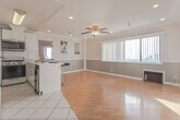 Located in the Hollywood Hills neighborhood of Los Angeles, this single-floor residence at 2069 Argyle Ave offers a thoughtfully designed layout with a bright, open feel throughout. The main living area features warm hardwood flooring, crown molding, and a ceiling fan with light fixture. Large windows along one wall flood the space with natural light while framing views of the surrounding cityscape, creating an airy and inviting atmosphere. The two-tone gray wall treatment adds a contemporary touch to the living area.

The kitchen is efficiently designed with white cabinetry, white countertops, and a suite of appliances including a gas range, stainless steel dishwasher, microwave, and full-size refrigerator. A curved breakfast bar with barstool seating provides a casual dining option and connects the kitchen seamlessly to the living space. White tile flooring grounds the kitchen area and transitions naturally to the hardwood of the adjacent rooms.

The bedroom is generously proportioned, featuring the same hardwood flooring, crown molding, and a ceiling fan, with a window offering pleasant outdoor views including palm trees. The bathroom is finished with warm travertine tile throughout the shower/tub surround, complemented by granite countertop accents, lending a spa-like quality to the space.