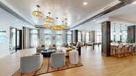Located at 904 Lake Park Blvd in Carolina Beach, NC, this single-story coastal clubhouse offers a generously sized, open-concept layout designed with a refined beach-inspired aesthetic. The main living area is anchored by multiple seating arrangements — featuring neutral sofas, soft blue accent chairs, and layered woven rugs — all beneath a striking tongue-and-groove ceiling adorned with bold reclaimed wood beams and cascading rattan pendant lights. Light wood-toned flooring flows seamlessly throughout, reinforcing the airy, coastal atmosphere. Large glass doors and tall windows line one wall, flooding the interior with natural light and offering direct views of the outdoor pool area. Rustic wood columns punctuate the open floor plan, adding warmth and architectural character. The game area includes a pool table and shuffleboard, providing recreational amenities within the space. A bar and counter seating area features rattan-backed stools set against a dramatic tropical botanical wallcovering, with a beverage station nearby. Coastal-themed artwork and decorative wall accents contribute to the cohesive design narrative throughout. Additional flexible space is accessible through black-framed glass doors, suitable for meetings or events. This clubhouse is a well-appointed communal space that balances sophisticated coastal design with functional, social amenities.