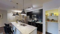 This single-floor space showcases a thoughtfully designed interior with a strong emphasis on modern aesthetics and functional living. The kitchen serves as the centerpiece, featuring striking dark cabinetry that extends to the ceiling, offering generous storage alongside a built-in wine rack. Gleaming granite countertops provide ample prep space and extend seamlessly across a large central island, which is equipped with an undermount stainless steel sink. Three bold industrial-style pendant lights hang above the island, adding both character and focused task lighting. The kitchen is fully equipped with stainless steel appliances, including a French door refrigerator, electric range, and built-in microwave, all complemented by a shimmering mosaic tile backsplash that adds texture and depth. Track lighting along the ceiling ensures the space is well-lit throughout. Rich, dark hardwood flooring runs through the area, grounding the design with warmth and contrast. A dining area is visible just beyond the kitchen, offering a natural flow between spaces. The overall ambiance is contemporary and polished, balancing dark, dramatic tones with bright surfaces to create an inviting yet sophisticated environment.
