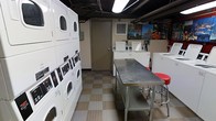 This single-floor laundry facility is designed with practicality and efficiency in mind. The space is outfitted with a row of stacked front-load dryers along one wall, maximizing vertical space to accommodate multiple units within a compact footprint. Complementing these are standalone top-load washing machines positioned along the opposite side, offering users a variety of laundry options. At the center of the room sits a stainless steel folding table, providing a sturdy and easy-to-clean surface for sorting and folding laundry. A pair of red stools nearby offers convenient seating while waiting. The flooring features a checkered tile pattern in neutral tones, contributing to the clean and utilitarian aesthetic of the space. Overhead, fluorescent strip lighting ensures the area is well-lit throughout. The walls are accented with colorful photographic prints, adding a touch of visual interest to the otherwise functional environment. A wall-mounted HVAC unit helps maintain a comfortable temperature. The space also includes a utility door at the rear, likely providing access to storage or mechanical areas. Overall, the layout is thoughtfully organized to support efficient, high-volume laundry use.