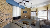 This single-floor space is a well-appointed communal lounge designed for both relaxation and social interaction. The interior is anchored by a striking curved brick fireplace that serves as a dramatic focal point, seamlessly integrated into the rounded exposed brick wall that gives the space a warm, rustic character. The seating area features comfortable upholstered sofas arranged around low-profile coffee tables, creating an inviting atmosphere for casual gatherings.

A flat-screen television is mounted directly on the brick wall, enhancing the entertainment functionality of the lounge. Ceiling fans are installed throughout, contributing to comfort and airflow across the space. The flooring is covered in a neutral-toned carpet that complements the warm tones of the brick.

Adjacent to the main lounge area is a bar-height dining section outfitted with sleek metal stools and a counter, offering an alternative seating arrangement. Windows dressed with horizontal blinds allow natural light to filter in while maintaining privacy. The ceiling treatment includes a combination of white paneling and recessed lighting, adding a polished contrast to the rustic brick elements. Overall, the space strikes a balance between industrial charm and contemporary comfort, making it a functional and welcoming environment.