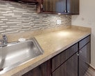 This single-floor residential unit offers a well-organized layout that balances comfort and practicality. The kitchen is a standout feature, equipped with rich dark wood cabinetry, light stone-toned countertops, a stainless steel sink, and a stylish linear mosaic tile backsplash in neutral tones of gray and beige. Under-cabinet lighting highlights the workspace, and the kitchen connects seamlessly to the adjacent living areas. The living room is generously proportioned, finished with wall-to-wall neutral carpeting and bright white walls that maximize the sense of light and space. A ceiling vent provides climate control, and the room opens visually toward the kitchen through an arched passageway. A separate dining or flex area features a pendant light fixture and additional carpeted space. The bedroom is a comfortable, carpet-floored room with a large window dressed in vertical blinds that allow natural light to filter in. It includes a built-in closet with shelving and bifold doors, offering practical storage. The bathroom is cleanly finished with white subway-style wall tiles, accented by a decorative mosaic border in warm brown and gray tones, and includes a full soaking tub. Throughout, the unit maintains a clean, neutral aesthetic that provides a versatile canvas for personalization.