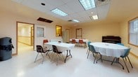 Parks Edge Community Room is a well-appointed, single-floor community space designed to accommodate a variety of gatherings and events. The main hall is a generously sized, open room featuring a drop ceiling with recessed fluorescent lighting and large skylight panels that flood the space with bright, even illumination. Multiple windows fitted with horizontal blinds line one wall, offering views of the surrounding outdoor greenery while maintaining a light and welcoming ambiance. The room is furnished with folding tables dressed in white linens and an assortment of chairs, providing flexible seating arrangements. A piano positioned along one wall adds a notable amenity for events requiring musical accompaniment.

An adjoining children's area offers a dedicated space equipped with child-sized tables and colorful seating, along with extensive built-in cabinetry for organized storage of supplies and equipment.

The kitchen is a functional and well-stocked space featuring oak cabinetry, ample countertop workspace, a dishwasher, microwave, coffee maker, and a utility sink—making it fully capable of supporting catered events or light meal preparation.

Throughout, the space maintains warm, neutral tones with wood-trimmed doorways, creating a practical yet inviting environment suited for community use.