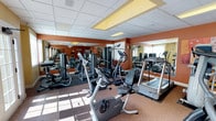 This fitness center offers a comprehensive workout environment thoughtfully designed to accommodate a variety of exercise routines. The space is outfitted with an impressive selection of Precor cardio equipment, including treadmills, elliptical trainers, and stationary bikes, arranged to allow comfortable movement throughout the floor. Complementing the cardio area is a dedicated strength training section featuring a variety of cable and weight machines suited for full-body conditioning. Large mirrors line the walls, visually expanding the space and allowing users to monitor their form during workouts. The drop ceiling with recessed lighting panels ensures the room is evenly and brightly illuminated, while natural light filters in through windows along one wall, enhancing the overall ambiance. The flooring consists of a durable, cushioned carpet suited for gym use, providing comfort underfoot. The warm, terracotta-toned accent walls add a welcoming energy to the environment. Overall, the fitness center presents a clean, organized, and fully functional space that caters to both casual exercisers and dedicated fitness enthusiasts.