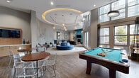 This thoughtfully designed amenity lounge offers a sophisticated and welcoming environment across a single expansive floor. The space is anchored by a dramatic circular recessed ceiling with warm ambient lighting, creating a focal point above a round, blue sectional sofa at the heart of the room. Soaring double-height ceilings and oversized windows flood the interior with natural light, offering views of the surrounding urban landscape. Wood-look flooring runs throughout, complementing a neutral grey and teal color palette accented with bold blue furnishings.

The lounge is divided into several distinct zones. A billiards area features a full-size pool table, while a stylish bar counter with a marble-look surface and industrial bar stools sits beneath pendant lighting and a wall-mounted television. Teal leather booth seating provides an intimate dining or gathering nook, adorned with framed artwork. A separate seating area hosts cushioned lounge chairs arranged around a circular ottoman, beneath decorative metallic wall sculptures. A round dining table with upholstered chairs sits beside a large window. French doors open to an outdoor terrace, enhancing the connection between interior and exterior. The overall ambiance is modern, vibrant, and well-appointed for social gatherings.