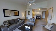 This single-floor residence showcases an open-concept layout that seamlessly connects the living area to the kitchen, creating a sense of spaciousness and ease of movement. The living room features notably high ceilings, enhancing the airy atmosphere, and is equipped with a ceiling fan that provides both functional ventilation and a subtle design element. Neutral-toned walls complement the light beige carpeting underfoot, establishing a warm and cohesive aesthetic throughout the space.

The living area is arranged around a dark-finished rectangular coffee table, flanked by a full-sized sofa and a generously sized chaise-style chair, both upholstered in charcoal gray fabric with patterned accent pillows. A large framed mirror on one wall adds depth, while a floor lamp provides soft ambient lighting. Decorative candles on the coffee table contribute to the inviting atmosphere.

The open kitchen, visible from the living area, features light wood cabinetry, white appliances including a range, and under-cabinet lighting that highlights the counter workspace. A dark-finished bistro-height dining table with matching chairs sits at the kitchen's edge, serving as a practical and stylish dining area. A hallway visible in the background suggests additional rooms beyond the main living space.