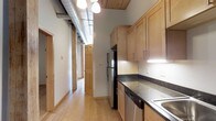 This single-floor space showcases a compelling blend of industrial and rustic design elements. The kitchen is a functional and visually striking space, featuring warm honey-toned wood cabinetry that extends to the ceiling, complemented by dark granite-style countertops and a stainless steel sink. Stainless steel appliances, including a refrigerator and dishwasher, add a modern touch. Overhead, pendant lighting provides focused illumination over the workspace.

One of the most distinctive architectural features is the exposed metal ductwork running along the ceiling, paired with corrugated metal ceiling panels and natural wood trim — a hallmark of loft-style industrial design. The light hardwood flooring flows seamlessly throughout, adding warmth and continuity across the space.

A long hallway extends from the kitchen, lined with classic wood-paneled doors that reinforce the natural material palette. The corridor creates a sense of depth and connects the various areas of the unit. The combination of raw industrial elements and warm wood tones gives the space a balanced, inviting ambiance suited to urban living.