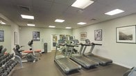 This fitness center is a dedicated, single-floor facility designed to support a comprehensive workout experience. The room features a drop ceiling with recessed panel lighting that provides bright, even illumination throughout the space. The walls are finished in a clean, neutral tone, contributing to an open and energizing atmosphere.

The cardio section is prominently featured, with a row of modern treadmills equipped with individual digital display consoles. Additional cardio equipment, including an elliptical trainer, is positioned nearby to offer variety. A weight bench with free weights is also visible, providing options for strength training.

The flooring consists of dark, durable gym-grade material suited for high-traffic athletic use. A water dispenser station is conveniently located along one wall, supporting hydration during workouts. Glass-panel doors at the rear of the room suggest access to an adjoining space, potentially offering additional equipment or amenities.

Decorative framed black-and-white sports photography adorns the walls, adding character and a motivational atmosphere to the environment. Overall, the fitness center is thoughtfully organized, clean, and functional, making it a practical and inviting space for daily exercise.