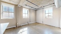 This single-floor loft-style unit showcases an industrial-chic aesthetic defined by polished concrete floors, raw exposed concrete ceilings, and visible HVAC ductwork throughout. The open kitchen serves as a central highlight, featuring warm wood cabinetry, stainless steel appliances including a full-size refrigerator, microwave, range, and dishwasher, along with a generous island with a clean white countertop and pendant lighting overhead — an ideal setup for cooking and entertaining. Track lighting runs along the ceiling, complementing the raw architectural character of the space. Multiple rooms benefit from large windows that invite abundant natural light, with views of the surrounding urban streetscape creating a connected city feel. A dedicated laundry area with a stacked washer and dryer adds practical convenience. The bathroom offers a double vanity with dark wood cabinetry, white countertops, and a spacious layout with additional storage. A bedroom includes a built-in closet with rod and shelving, accessed through a warm-toned wood door. Exposed concrete columns and white walls throughout balance the industrial elements with a clean, modern sensibility, making this a versatile and characterful urban living space.