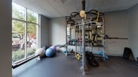 Located at 411 River St in Greenville, South Carolina, this single-floor fitness center offers a well-equipped and thoughtfully designed workout facility. The space features durable rubber flooring throughout, a drop ceiling with recessed lighting, and neutral-toned walls that create a clean, functional atmosphere.

The main workout area is divided into distinct zones to accommodate a variety of training styles. A cardio section houses multiple treadmills and an elliptical machine, positioned near warm yellow-accented walls and open floor space for stretching or bench work. Adjacent to this, a free weights area provides a full rack of dumbbells in varying sizes, accompanied by a workout bench, all bathed in abundant natural light streaming through large floor-to-ceiling windows that offer views of the surrounding greenery.

A dedicated functional training zone features a Life Fitness Synrgy360 multi-functional rig, complete with cable attachments, TRX suspension trainers, battle ropes, kettlebells, and stability balls — providing a comprehensive range of strength and conditioning options. The windows in this corner further enhance the space with natural light and outdoor views.

Near the entrance, a water station with bottle storage and a small refreshment stand ensures hydration convenience. Glass-panel sliding doors provide a clear sightline into the facility, contributing to an open and welcoming feel throughout the space.