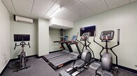 Located at 601 N Broad St in Philadelphia, PA, this fitness center offers a well-equipped exercise facility across a single floor. The space is thoughtfully organized to accommodate a variety of workout routines, with distinct zones for cardio, strength training, and functional fitness. The cardio area features multiple treadmills and elliptical machines, each equipped with individual digital consoles, complemented by a wall-mounted flat-screen TV for entertainment during workouts. The strength training section includes a full rack of dumbbells in varying weights alongside several selectorized resistance machines, providing options for both isolated and compound exercises. A dedicated functional fitness zone features stability balls, medicine balls, and a stretching board, anchored by a large floor-to-ceiling mirror that amplifies the sense of space. Throughout the facility, durable rubber-composite flooring provides comfort and safety underfoot, while a drop ceiling with recessed fluorescent lighting ensures consistent, bright illumination. Soft sage-green walls create a calm, focused atmosphere. A wall-mounted split AC unit maintains comfortable climate control. A sanitation station near the entrance promotes cleanliness. The layout is efficient and accessible, making this a comprehensive fitness amenity suited for a full range of exercise needs.