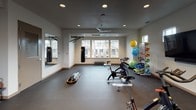 This fitness room is a comprehensive, single-floor exercise facility designed to support a wide range of workout routines. The space features durable dark flooring throughout, smooth neutral-toned walls, and a ceiling fitted with recessed lighting and a ceiling fan for comfort during intense sessions. Natural light fills the room through multiple windows, and a set of double glass doors opens directly to an outdoor balcony area, providing both ventilation and pleasant views of the surrounding exterior.

The room is outfitted with an impressive array of equipment. Cycling enthusiasts will find stationary spin bikes ready for use, while a wall-mounted flat-screen television provides entertainment or instructional content. A full dumbbell rack offers a range of free weight options for strength training, and a colorful medicine ball and stability ball rack adds versatility for functional fitness exercises. Two heavy punching bags are mounted along one mirrored wall, supported by a wall-mounted pull-up bar, creating a dedicated boxing and bodyweight training zone. Resistance sleds on the open floor allow for dynamic conditioning work. A large wall mirror enhances the sense of space and allows users to monitor their form. The room also includes a separate door leading to an adjacent area, rounding out this well-appointed and functional fitness facility.
