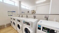 This shared laundry facility at 100 Dundas St E in Mississauga, Ontario, is a well-organized, single-floor space divided into two functional laundry rooms. The interior features light wood-look tile flooring throughout, white walls, and a clean, utilitarian design that prioritizes ease of use and efficiency.

The main laundry area is generously sized, housing multiple rows of commercial-grade front-loading washers and dryers with digital control panels. High-positioned windows allow natural light to filter in, contributing to a bright and airy atmosphere. Instructional signage for the washers and dryers is clearly posted on the walls for user reference. A stainless steel utility sink and a folding/sorting table provide practical workspace, while a payment kiosk offers a convenient cashless payment option for residents.

A secondary laundry room features additional top-loading and stacked washer-dryer units, with a floor drain centrally located for easy maintenance. Both rooms are equipped with safety features including a fire extinguisher and a security camera. A shared utility area connects the two laundry rooms, offering supplementary counter space, a seating area, and a waste bin. The facility is well-maintained and designed to serve the practical laundering needs of building residents efficiently.